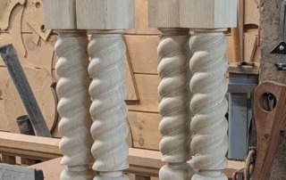 Four custom twisted white oak table legs with square tops and bases, displayed in a woodworking shop setting.