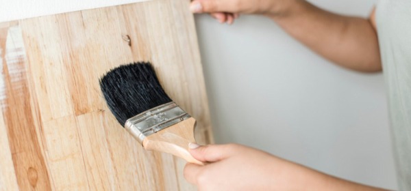 Man applying polyurethane to a dark-stained wood board with a wide brush.